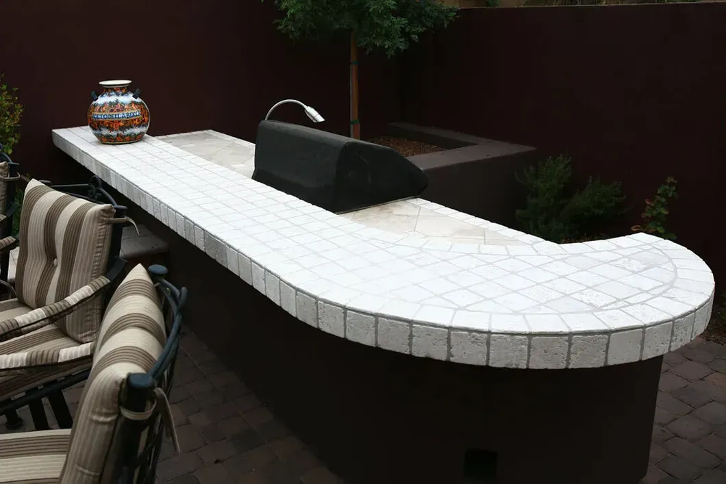 Outdoor kitchen with white tiled countertop, grill, and decorative vase. Dark brown base, and patio setting.