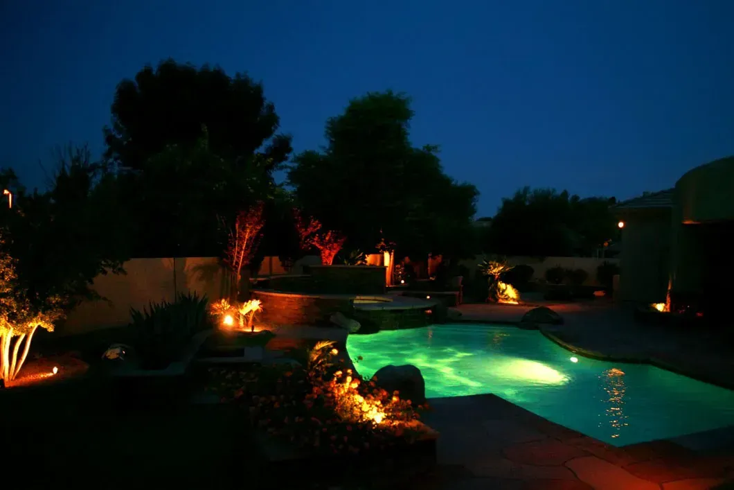 Nighttime view of a backyard with a lit pool, trees, and landscape lighting.