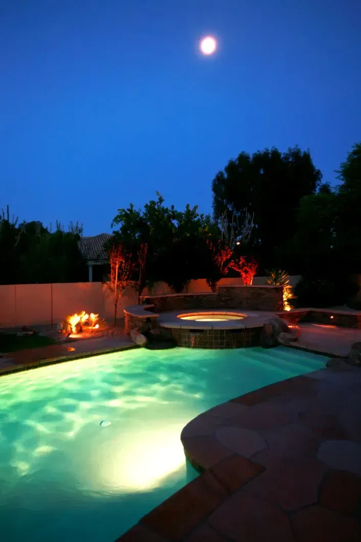 Night view of a swimming pool with a hot tub, under a full moon. Turquoise water illuminated by lights.