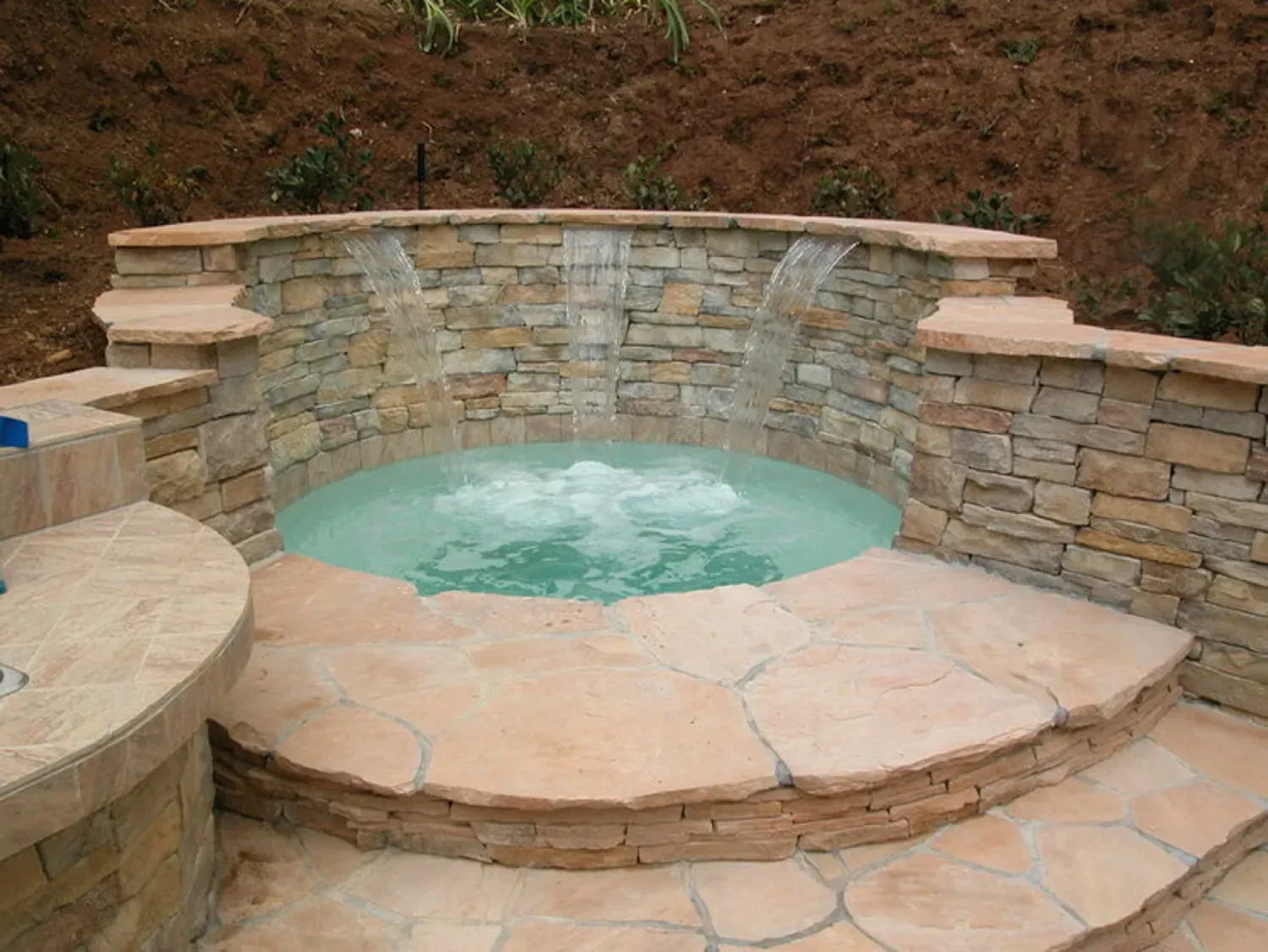 Stone-walled hot tub with waterfall feature, surrounded by stone patio.