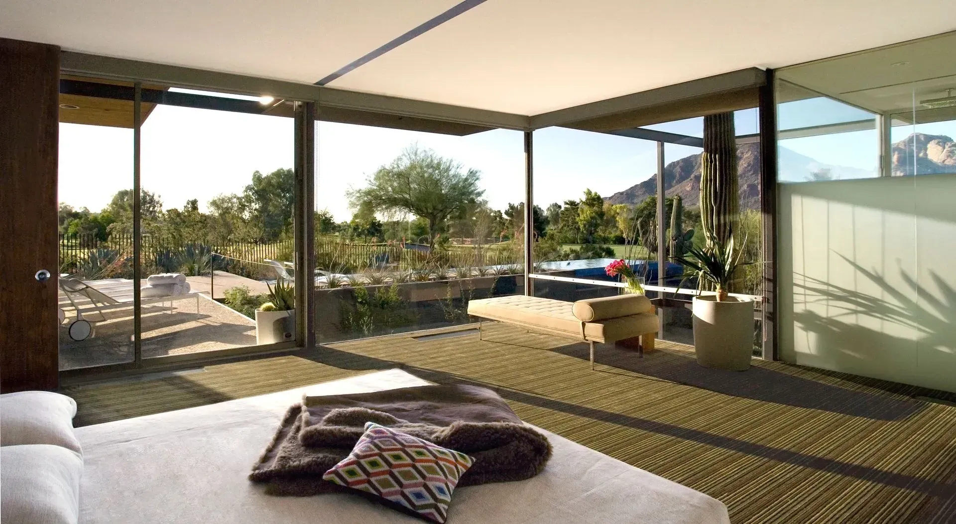 Bedroom with floor-to-ceiling windows overlooking a pool and desert landscape. Sunlight streams in.