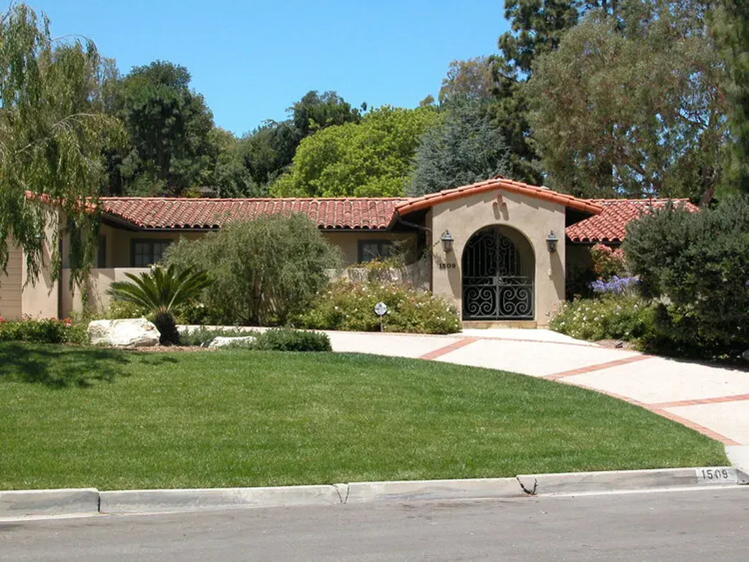 Tan house with red tile roof, arched entryway with decorative iron gate, green lawn and trees.