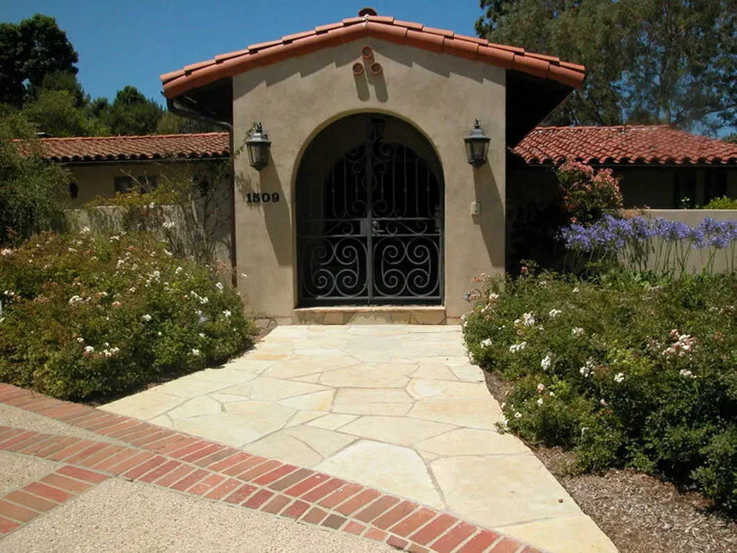Stone walkway leading to a tan stucco archway entrance with a wrought iron gate, flanked by flowering bushes.