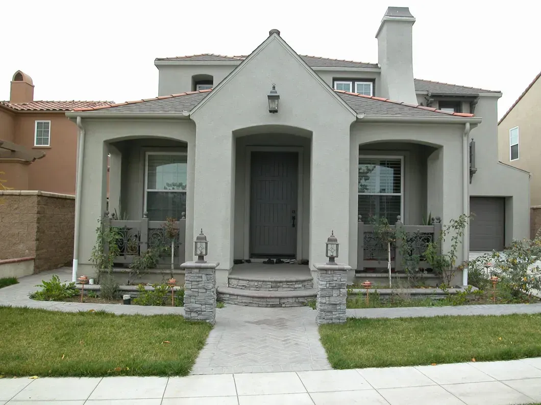 Two-story house with light gray stucco exterior, small porch, and a brick walkway leading to the front door.