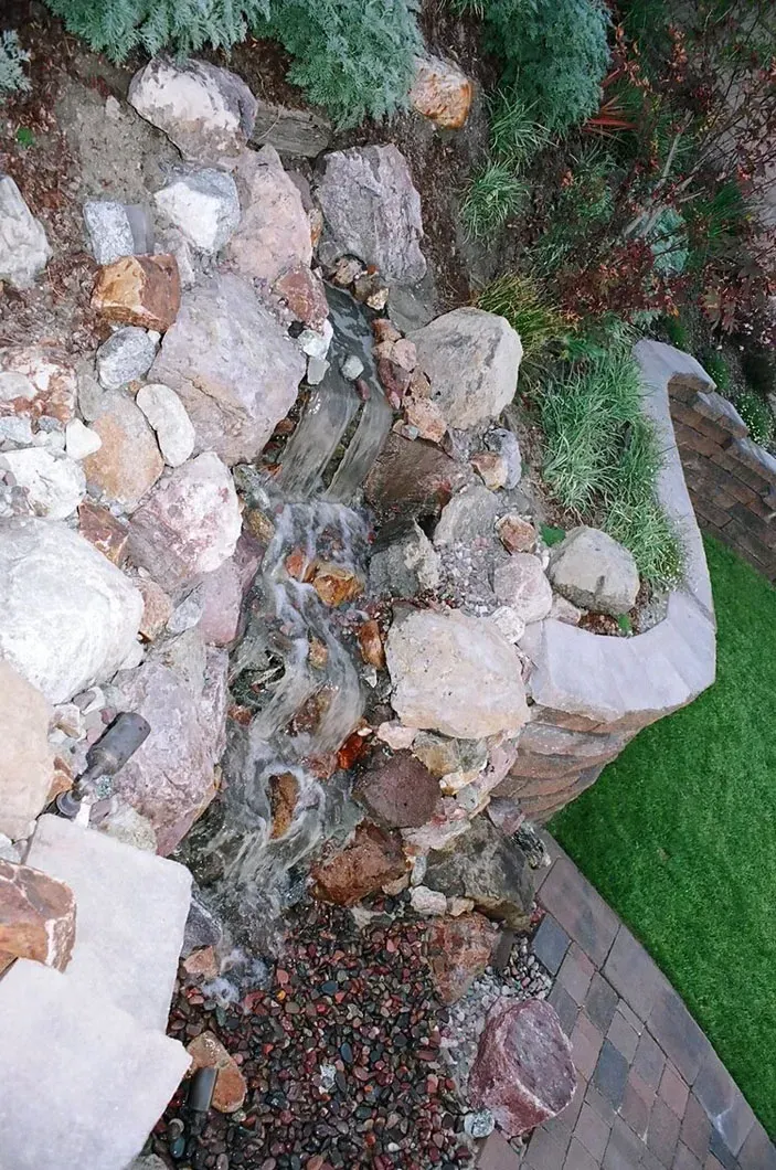 Water cascades over rocks, forming a small waterfall in a landscaped garden, with greenery and grass visible.