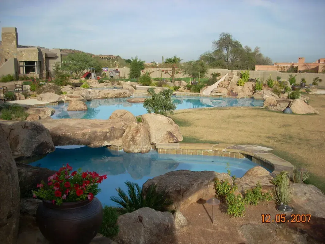 A stone-lined pool with blue water and rock formations, a building in the background, and dry landscape.