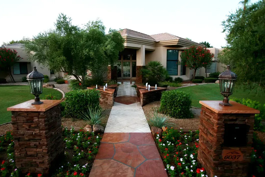 Stone pathway leading to a beige house, lined with bushes, flowers, and two lamp-topped columns.