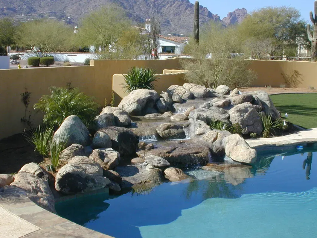 A rock waterfall feature flows into a blue swimming pool in a desert landscape.
