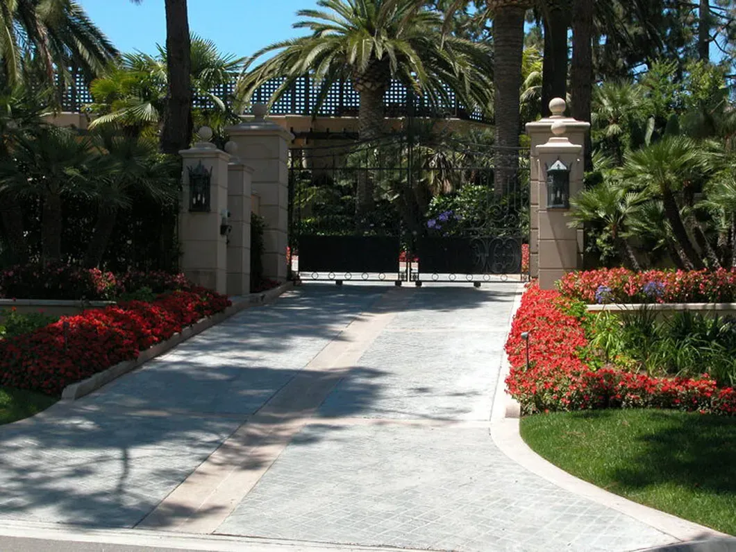 Gated driveway with stone pillars, manicured red flowers, and palm trees.
