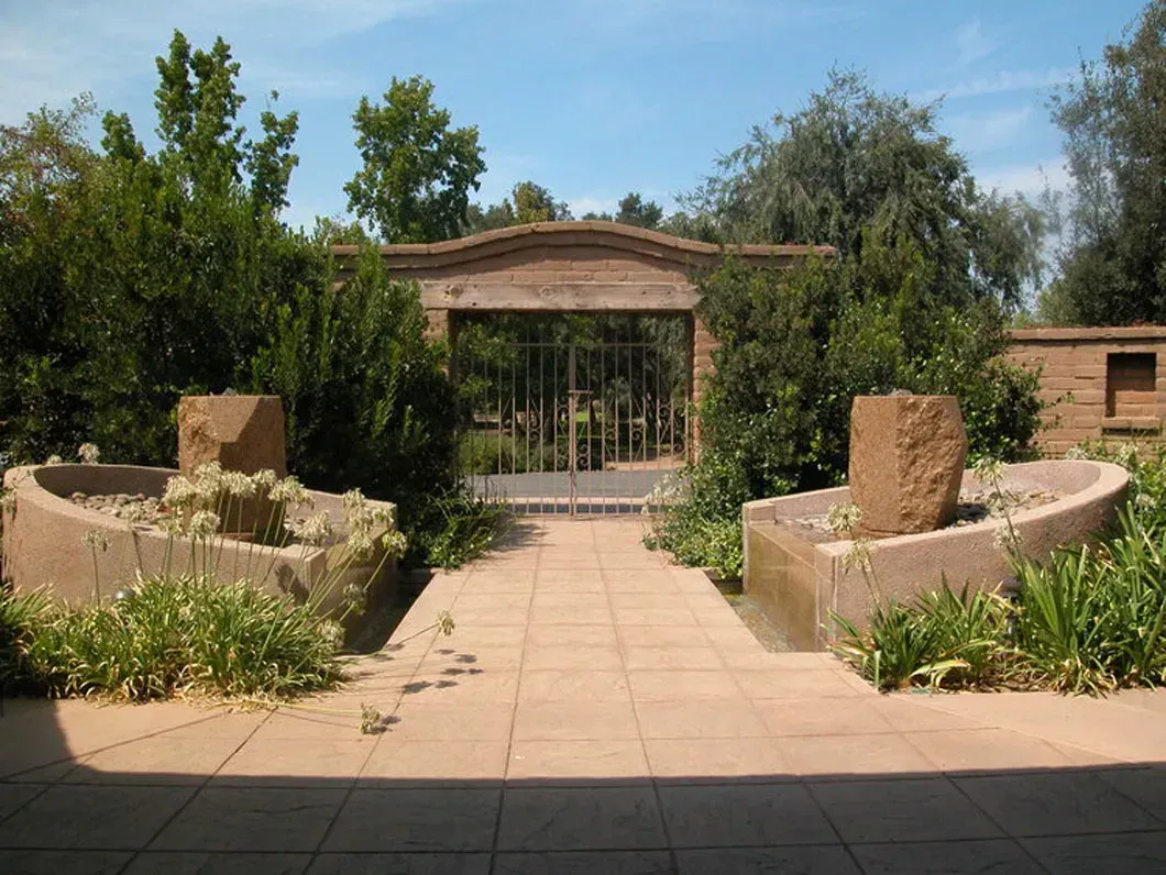 Stone patio with water features, leading to a gateway in a brick wall. Lush greenery surrounds the scene.
