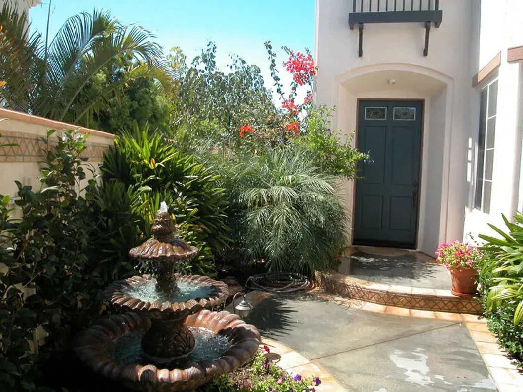 Fountain in a courtyard with lush greenery, leading to a front door of a white building.