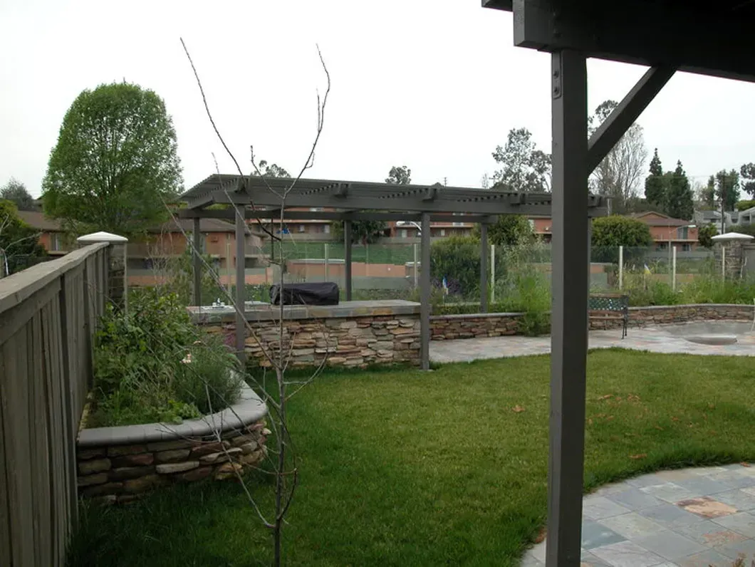 A backyard with a stone wall, pergola, and overgrown grass. View from a porch.