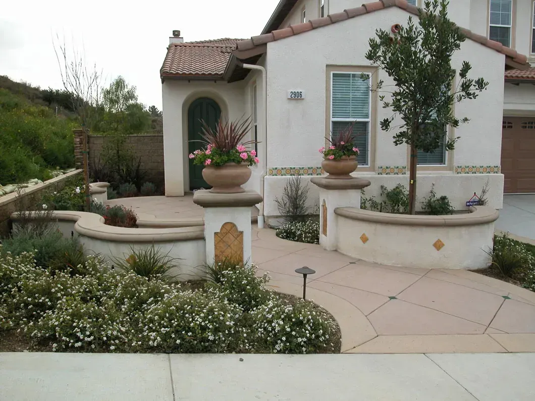 Spanish-style home with landscaped yard and pathway. Planters with flowers. Sunny day.