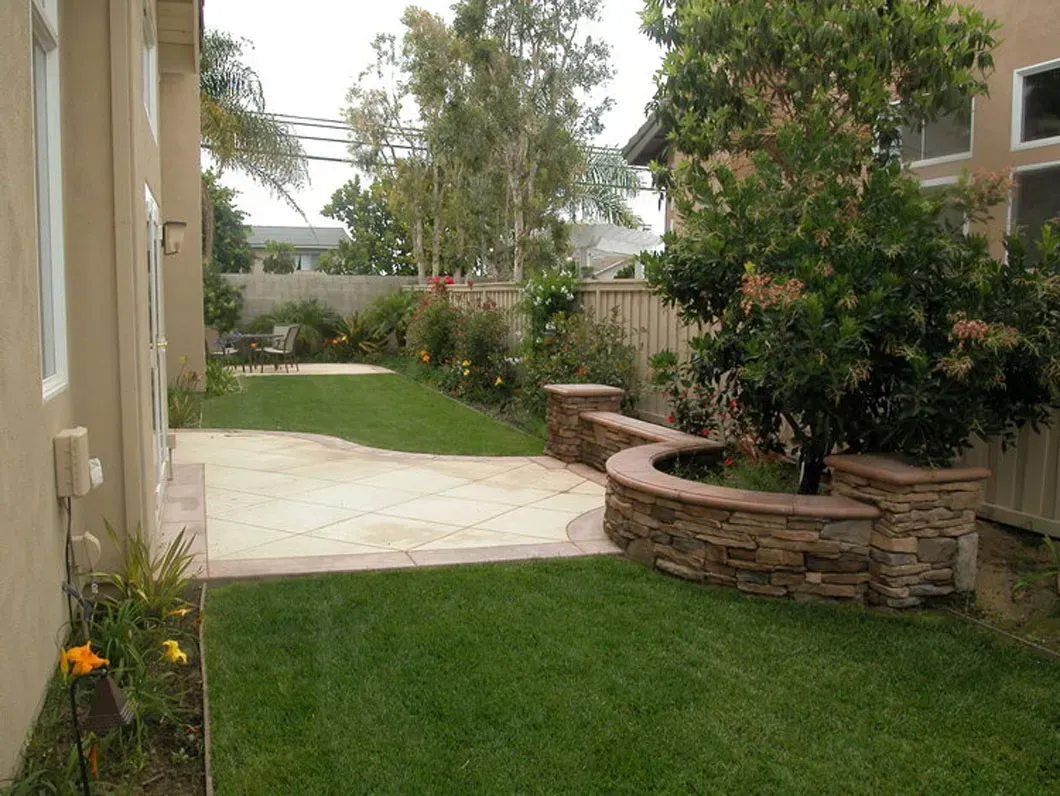 A backyard with a patio, lawn, and a stone-walled tree planter. Beige house siding on the left.