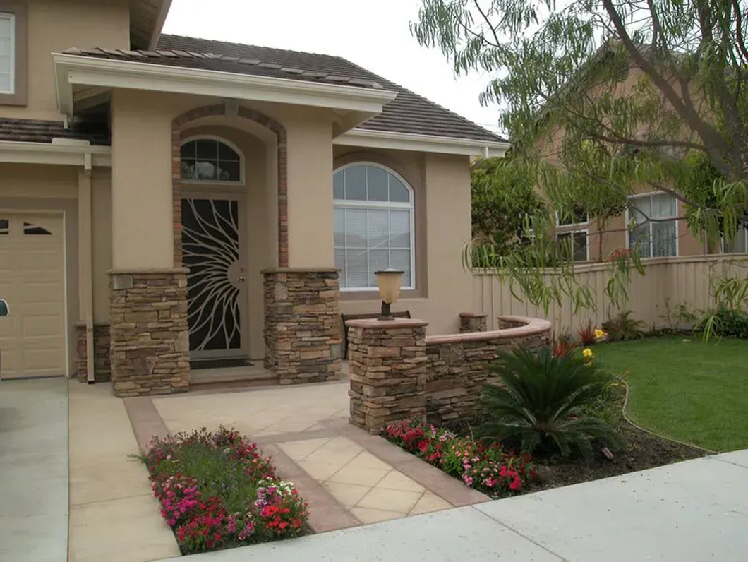Beige stucco house with stone accents, arched doorway, and flowerbeds along a walkway.