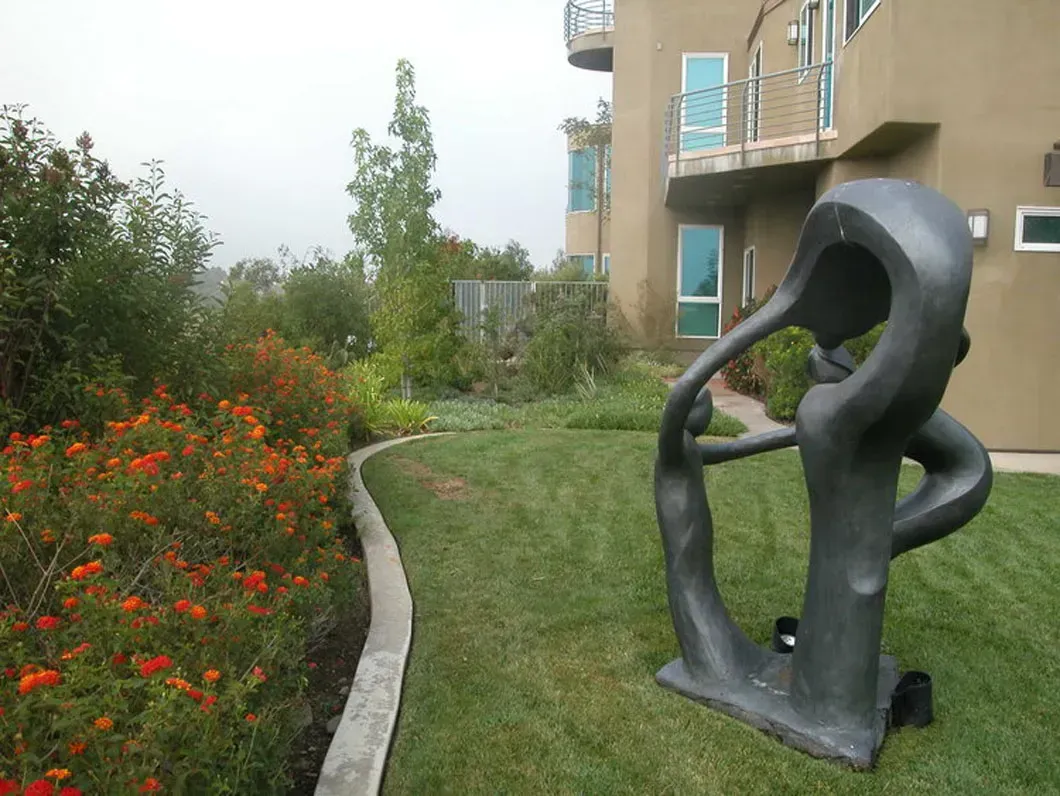 Sculpture on green lawn next to a building. Red flowers border a curved walkway, foggy day.