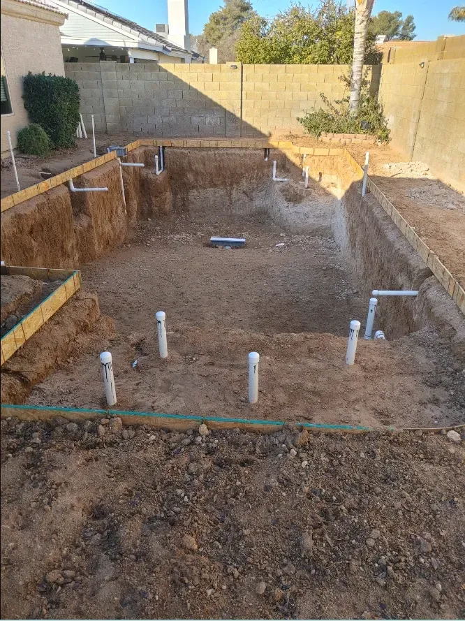 Pool excavation in a backyard, with exposed pipes and wood framing.