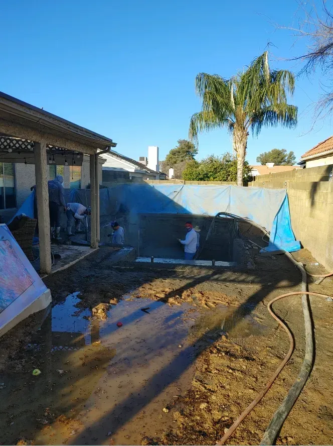 Construction workers renovating a backyard pool, covered in blue tarp. Muddy ground and palm tree are present.