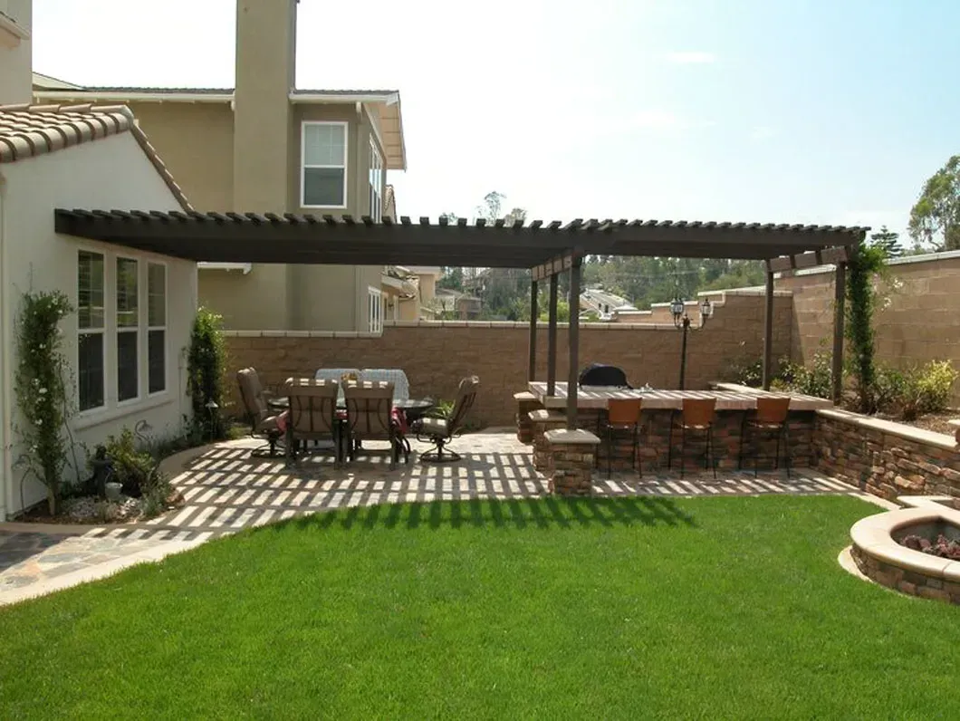 Lawn and patio with pergola, outdoor kitchen, dining table, and built-in fire pit. Sunny day.