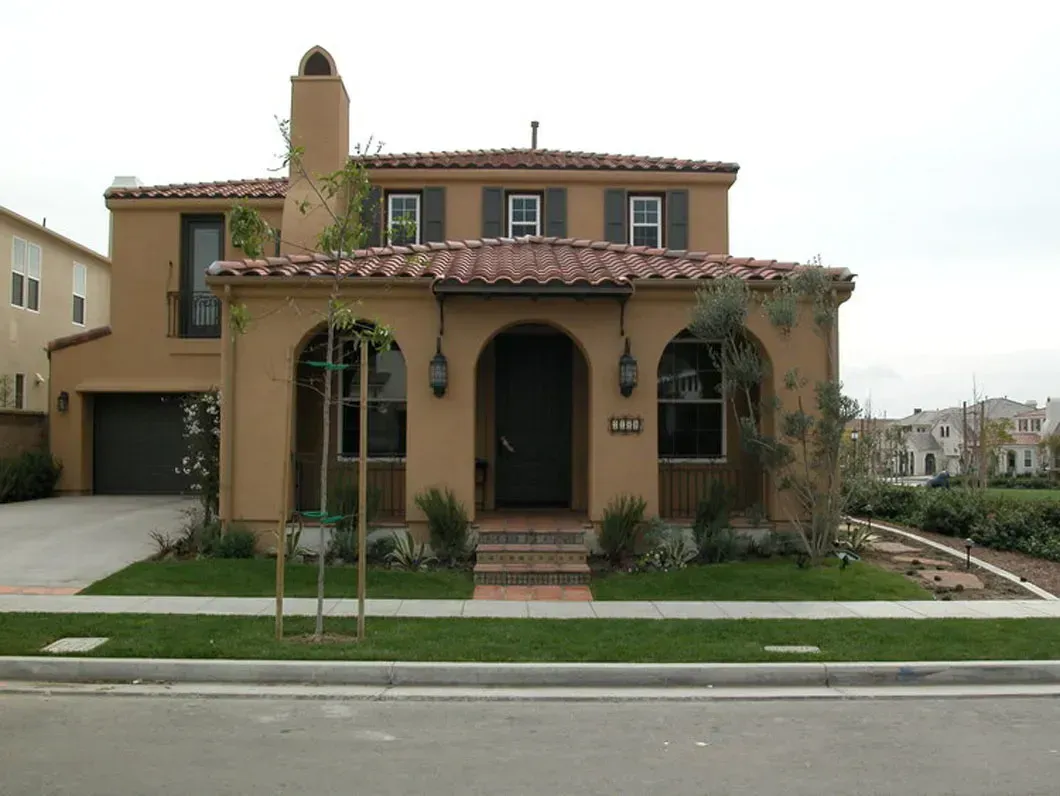 Two-story beige stucco house with a red tile roof and arched entry, with a green lawn.