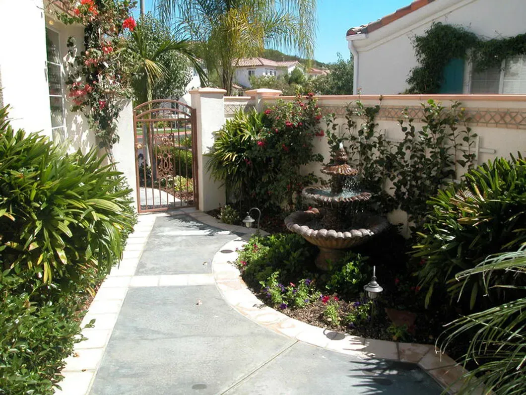 Path leading to wrought iron gate and a tiered fountain in a lush garden.