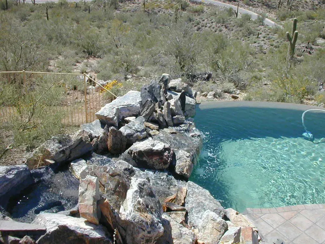 Waterfall feature next to a swimming pool, rocks and clear water, desert landscape in the background.