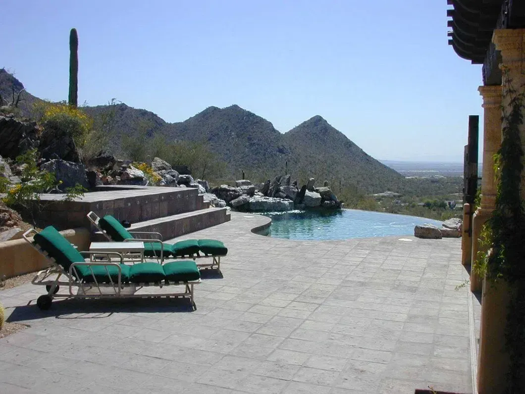 Lounge chairs beside an infinity pool overlooking desert mountains. Bright sunlight, clear blue sky.