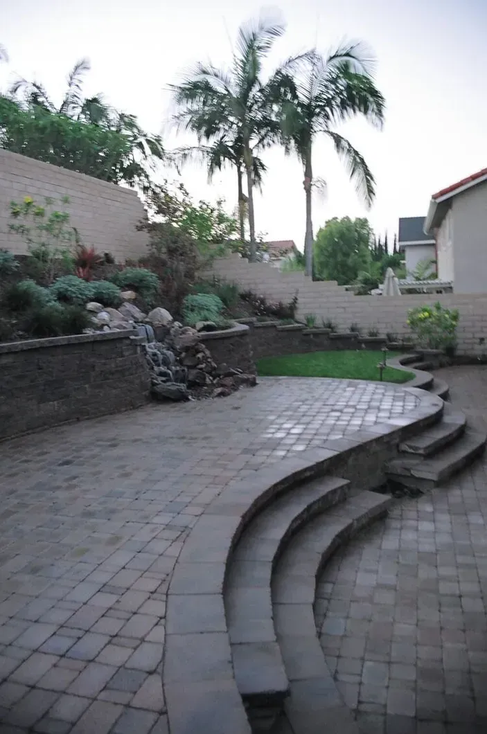 Backyard patio with tiered steps, waterfall, and stone walls; palm trees in background.