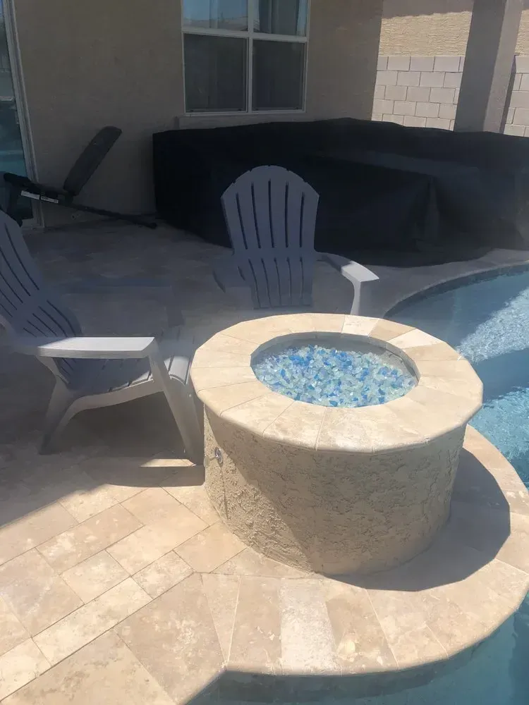 Stone fire pit with blue glass, two white chairs, pool, and covered hot tub on patio.