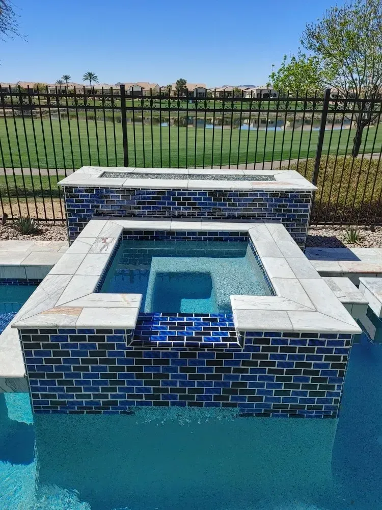 Swimming pool with a blue-tiled hot tub and a brick-lined fire pit, set against a lawn and fence.
