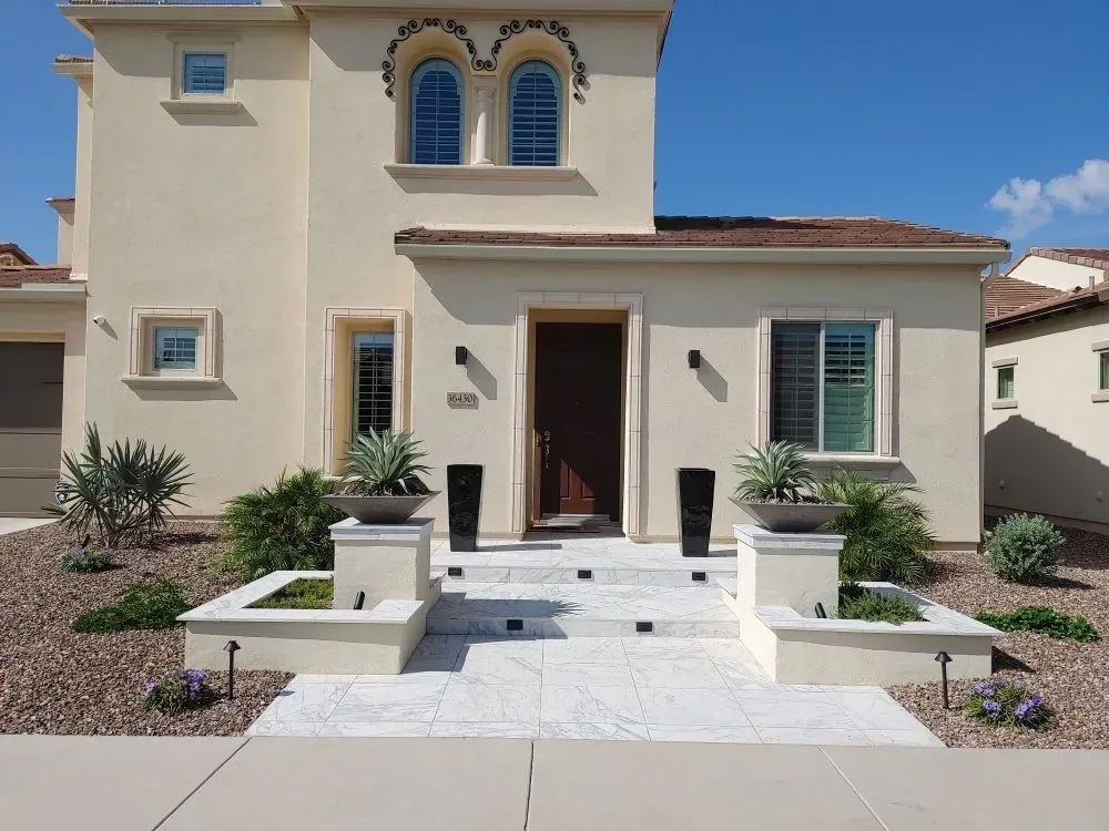 Beige two-story house with a brown door, stone steps, and decorative planters. Blue sky backdrop.