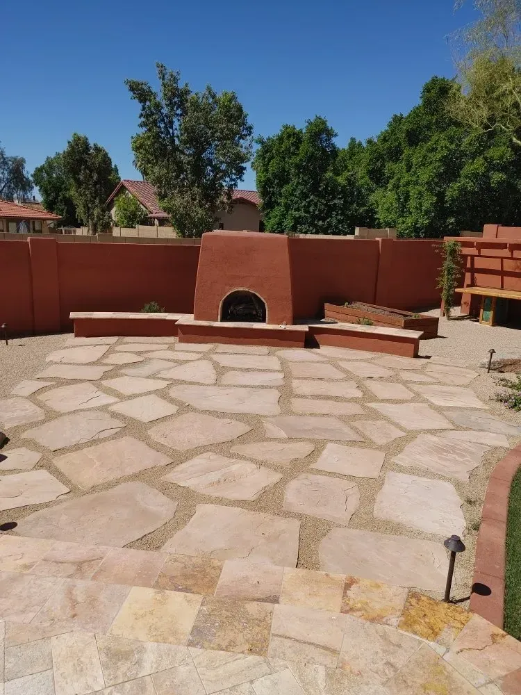 Patio with stone pavers, red stucco wall, fireplace, and desert landscaping under a blue sky.