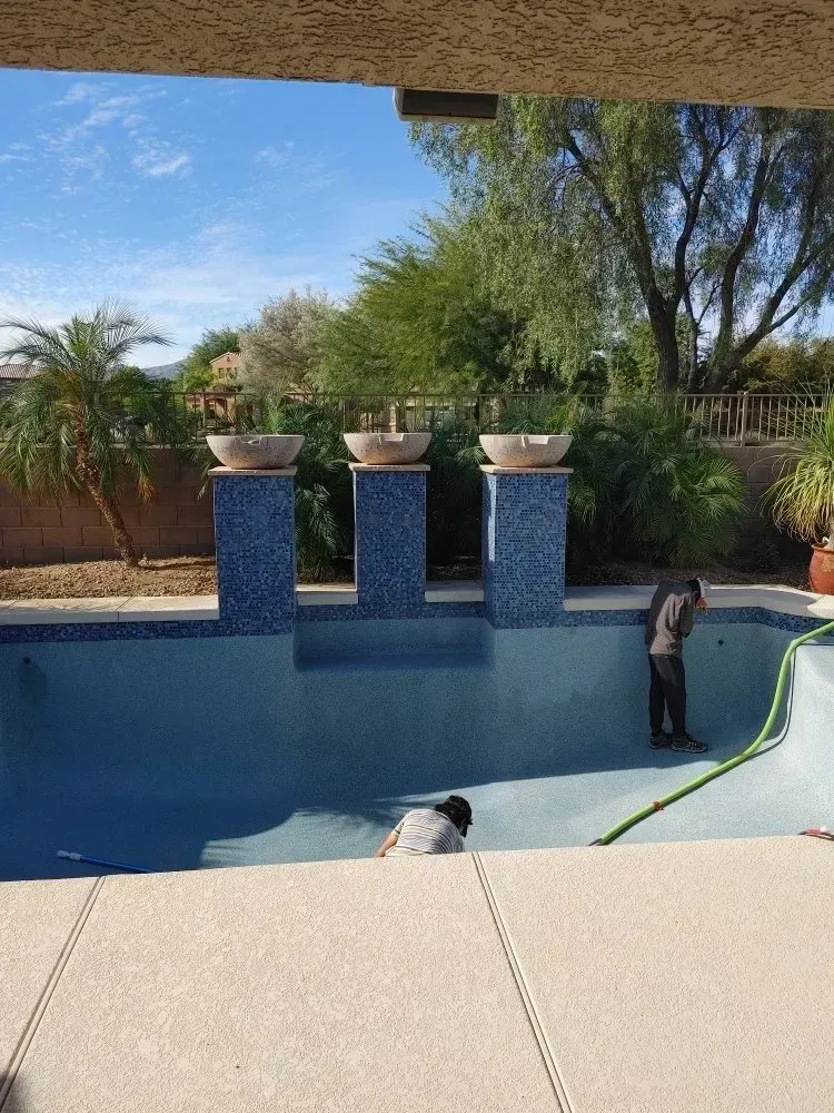 Workers cleaning a partially drained blue-tiled swimming pool. Three decorative pillars with bowls stand at the pool's edge.