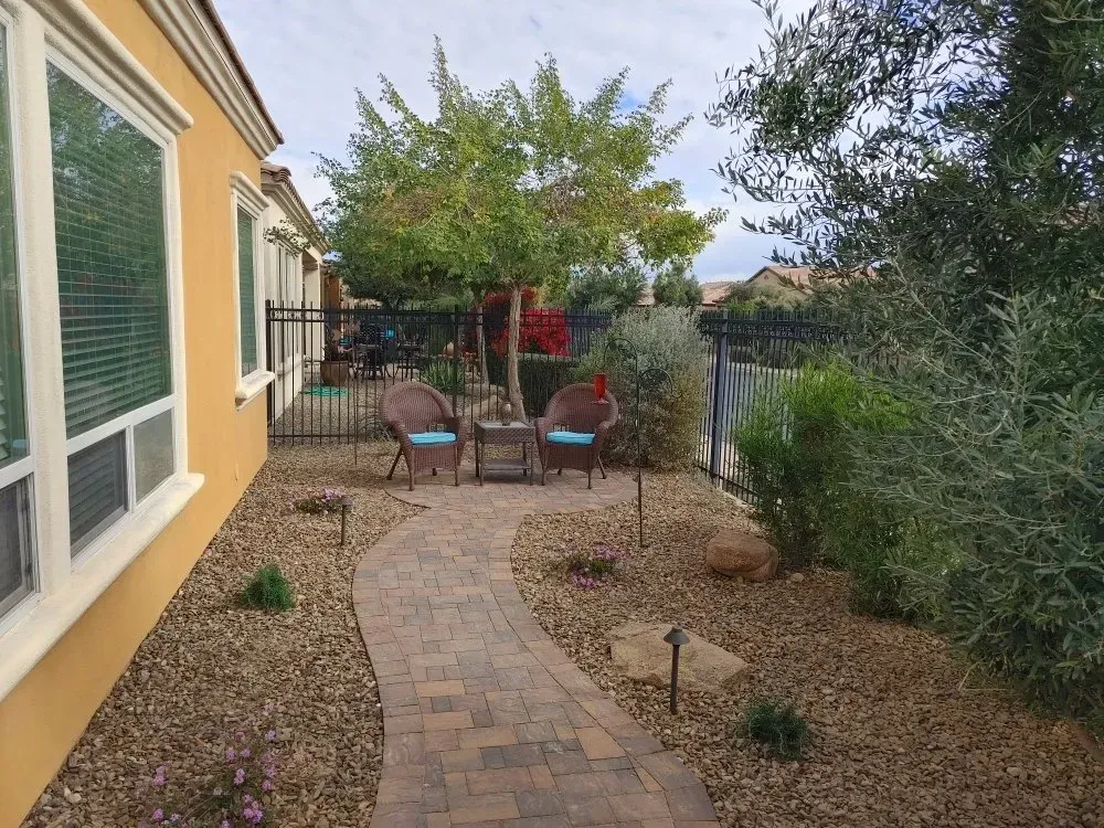 Brick pathway leads to a seating area with two chairs, next to a tree, within a fenced yard.