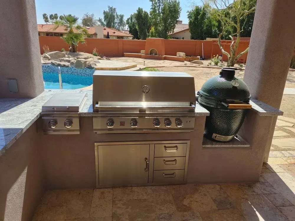 Outdoor kitchen with a stainless steel grill, a Big Green Egg, and built-in storage next to a pool.