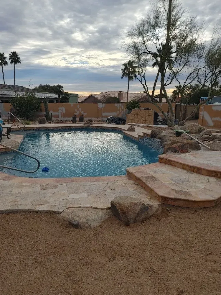 Pool with stone steps and surrounding sand, palm trees in the background, overcast sky.