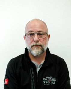 A man with a beard and glasses is wearing a jacket that says quality assurance roofing