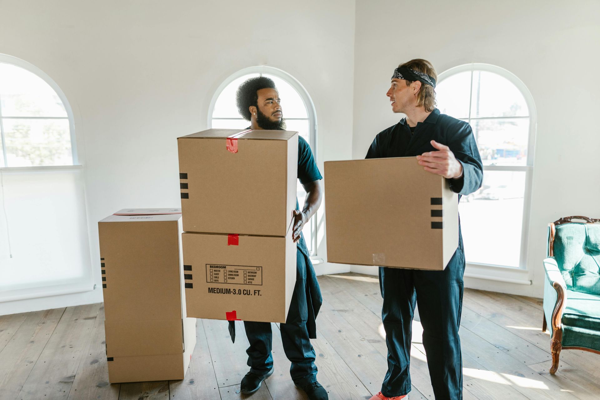 Two people move boxes in an empty room. One carries stacked boxes, the other a single box.