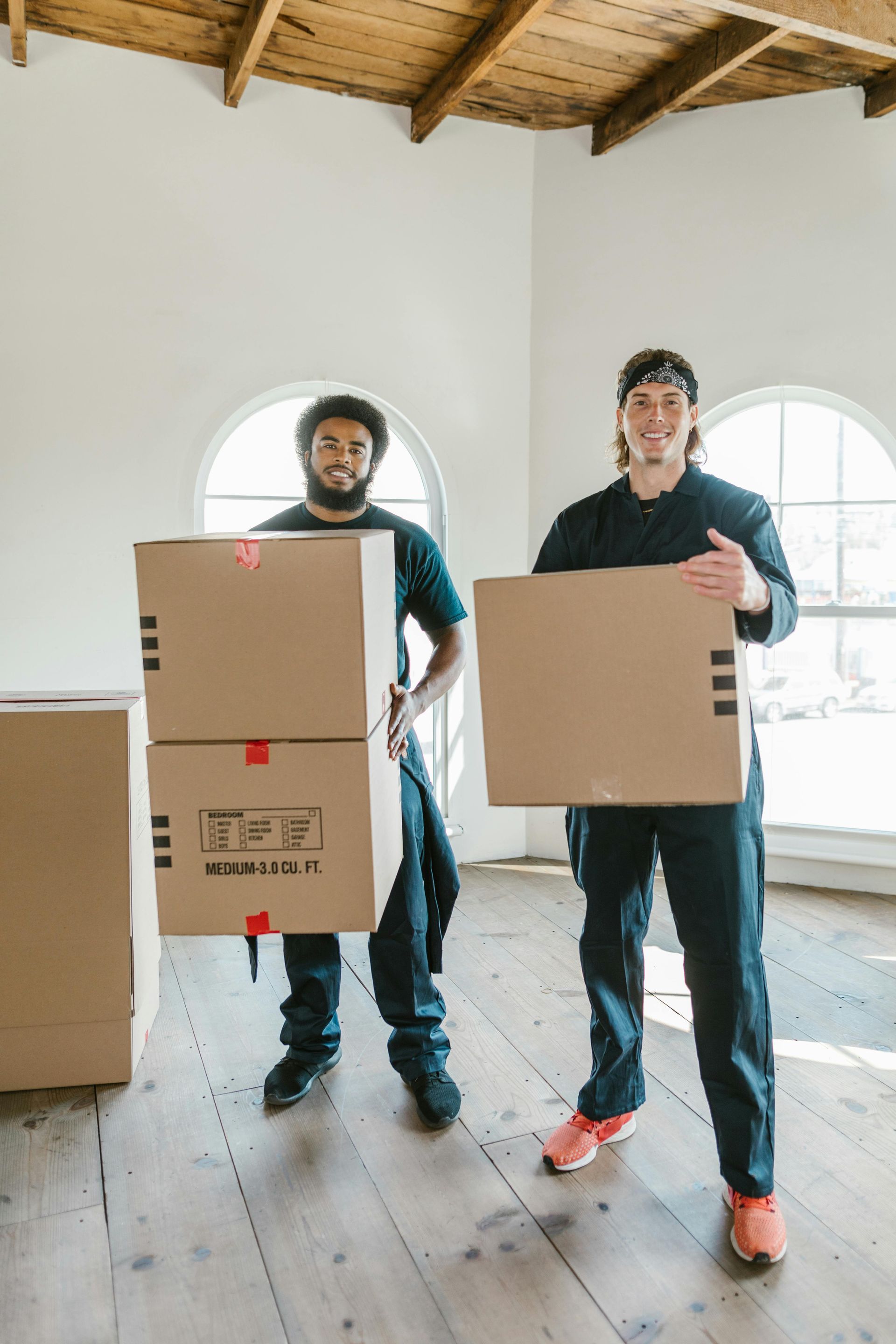 Two movers holding cardboard boxes in an empty room with arched windows and wood ceiling.