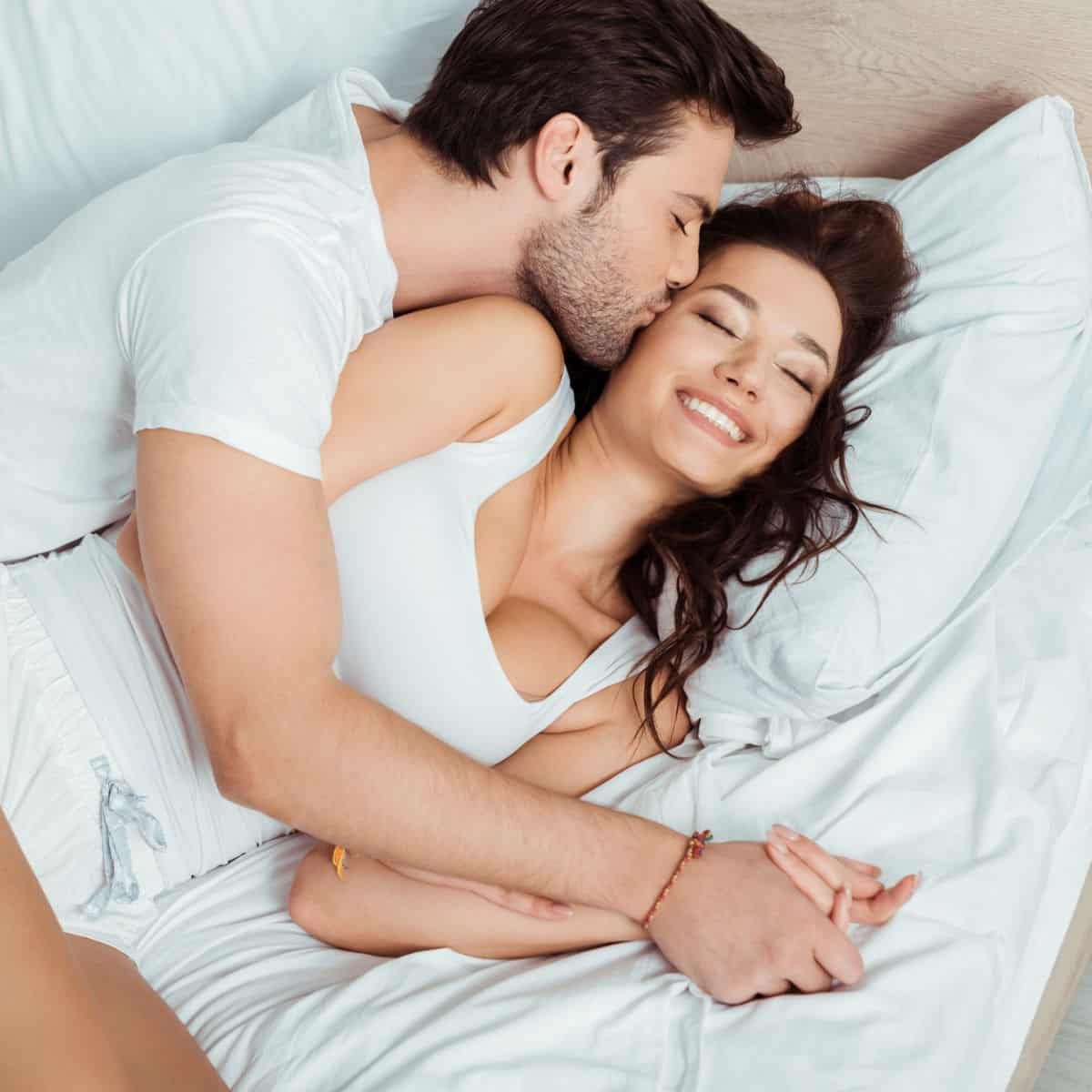 Couple cuddling in bed, smiling under white sheets and pillows
