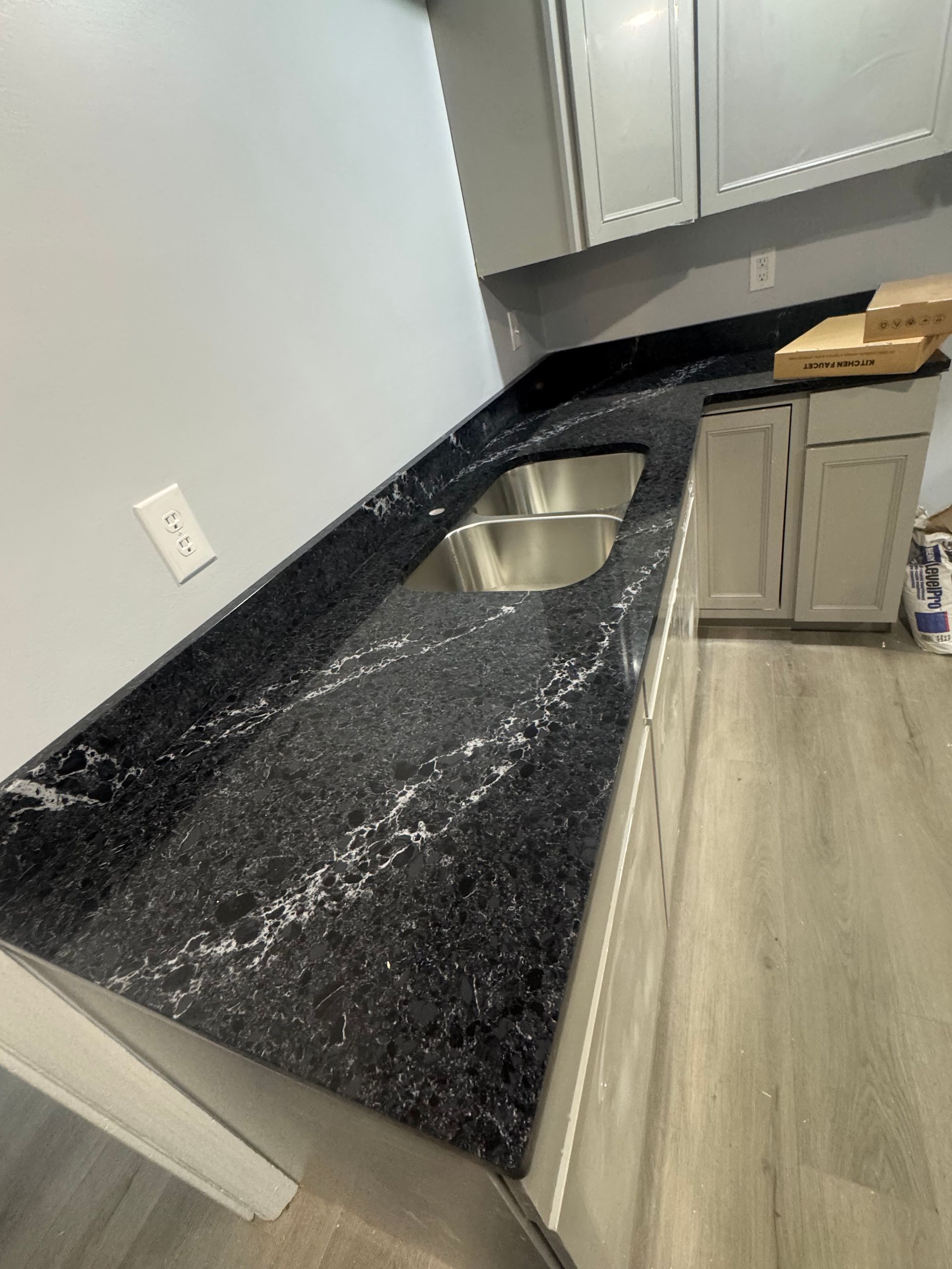 Black countertop with a stainless steel sink in a kitchen. Grey cabinets and light flooring.