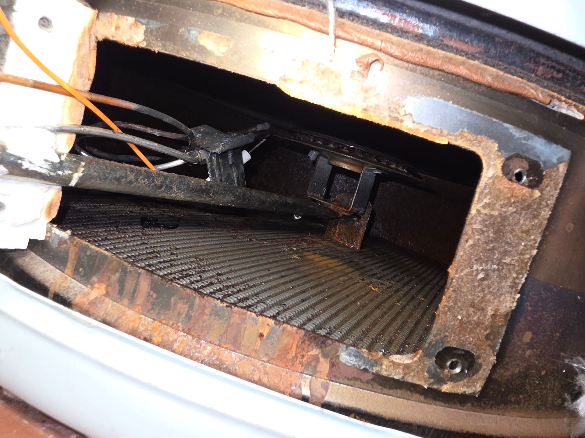 Interior view of a corroded metal furnace component. Wires and rust are visible within the dark space.