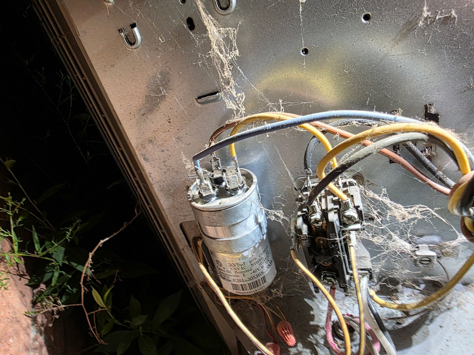 Close-up of AC unit wiring with capacitor and contactor, covered in cobwebs, outdoors.