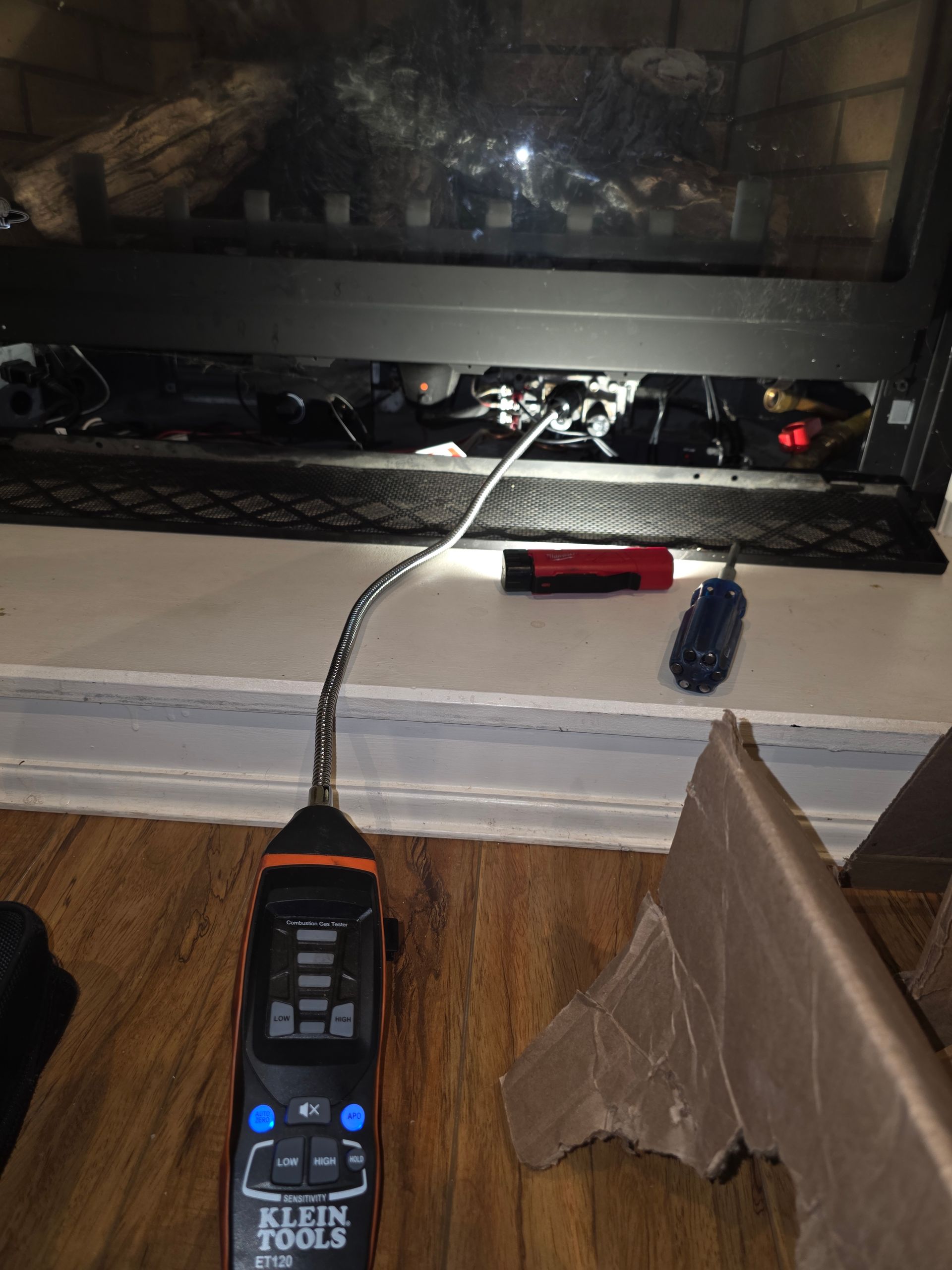 A black and orange Klein Tools electrical tester near a fireplace, cords, and a tool on a white mantel.