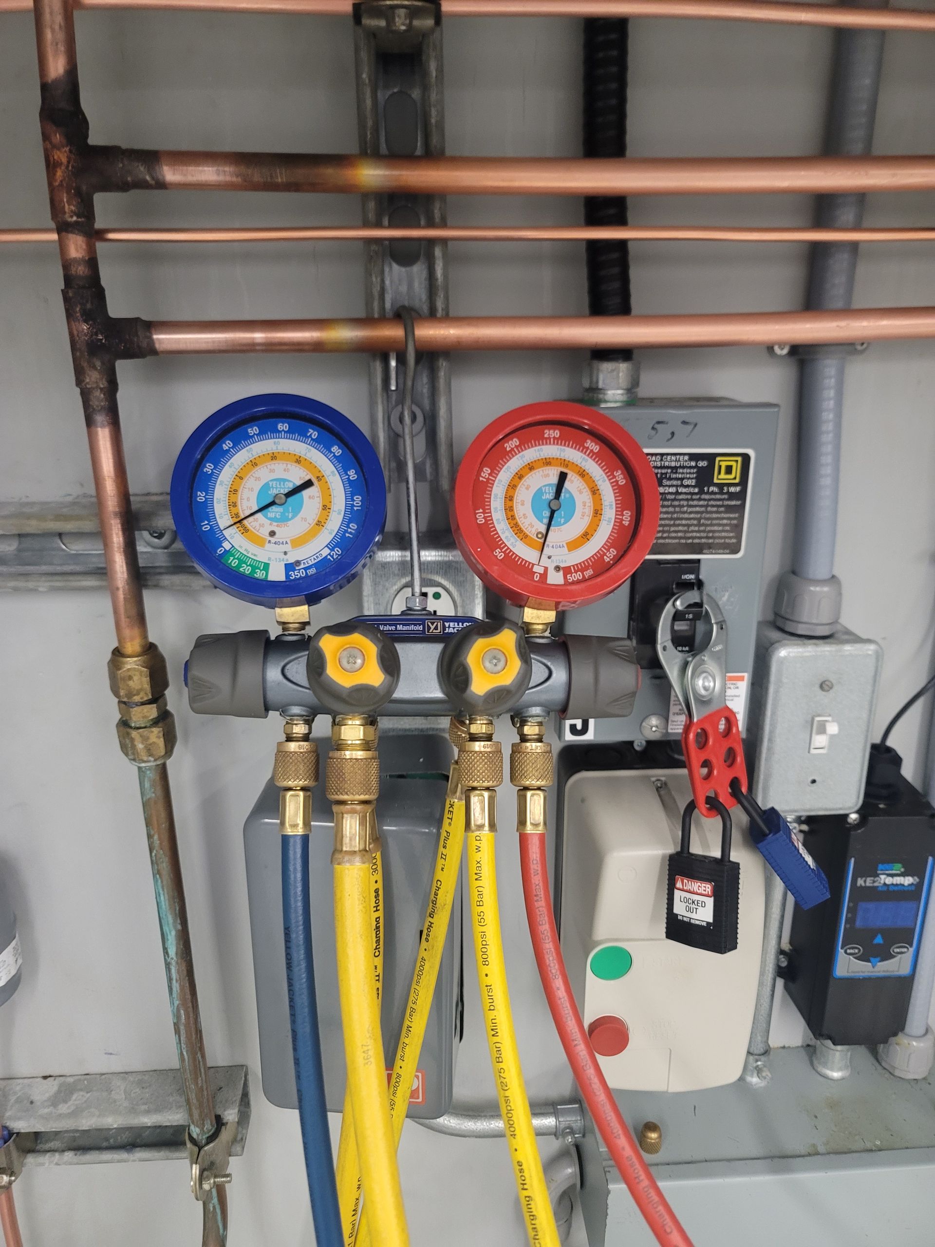Refrigeration gauges with blue and red dials, connected to yellow, blue, and red hoses, mounted on a wall with copper pipes.