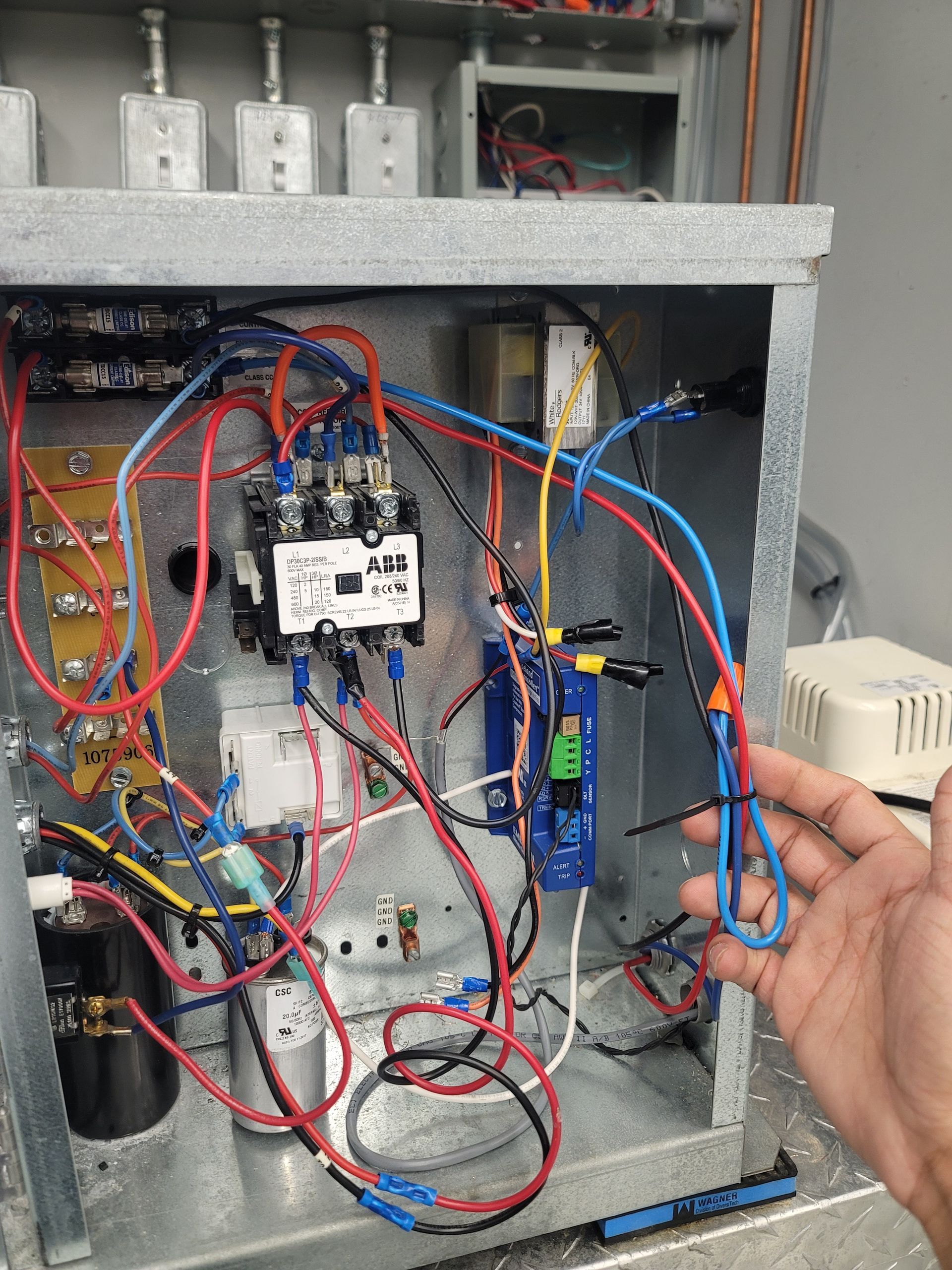 Hand holding a blue wire in an open electrical panel with various colored wires and components.