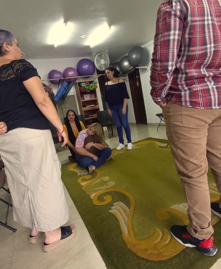 Personas en una sala con aparatos de ejercicio. Algunos observan a una persona sentada en una alfombra.