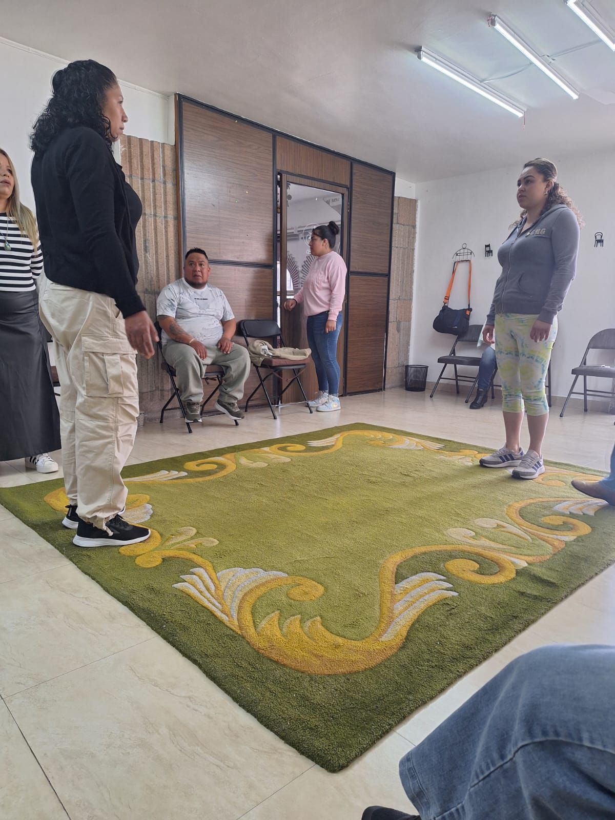 Personas en una habitación, sobre y alrededor de una alfombra verde. Algunos de pie, otros sentados.