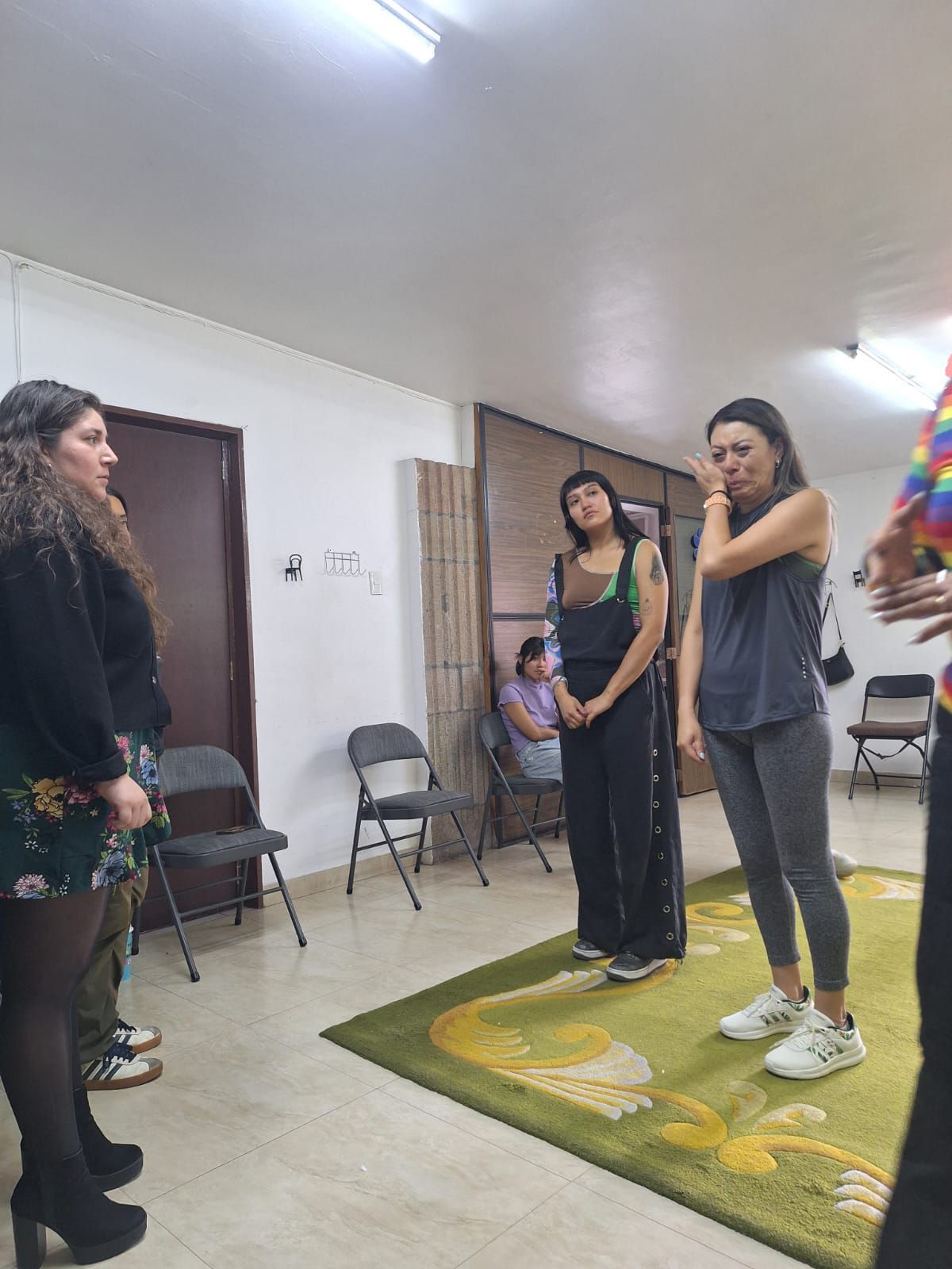 Varias personas en una habitación, algunas mirando, otras pareciendo emocionadas, de pie sobre una alfombra verde.