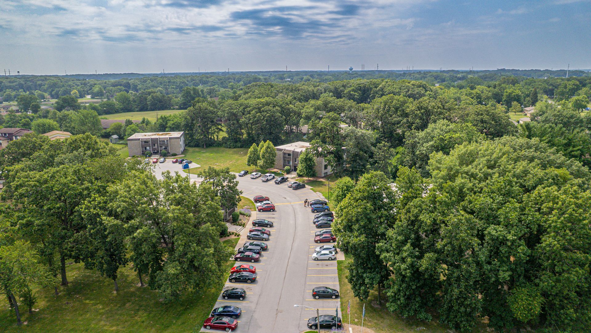 Aerial view of a parking lot lined with cars near apartment buildings, surrounded by dense trees and a distant skyline.